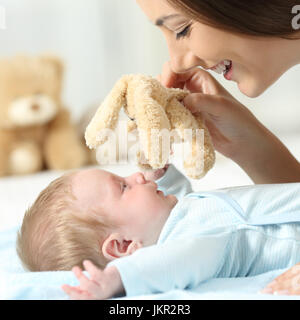 Side view close up portrait of a mother playing with her baby holding a teddy on a bed Stock Photo