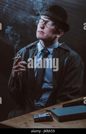 Young man sitting at table with gun while smoking cigarette Stock Photo