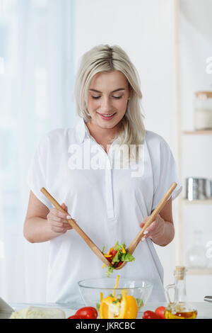 Smiling young woman tossing fresh salad on kitchen counter at home Stock Photo