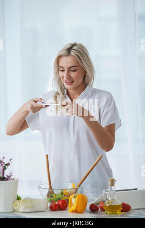 Smiling beautiful woman photographing salad through smart phone at kitchen counter Stock Photo
