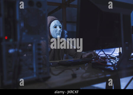 Man wearing mask and hood using computer at table Stock Photo