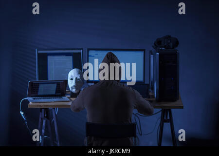 Rear view of computer hacker sitting at desk with equipment against gray background Stock Photo