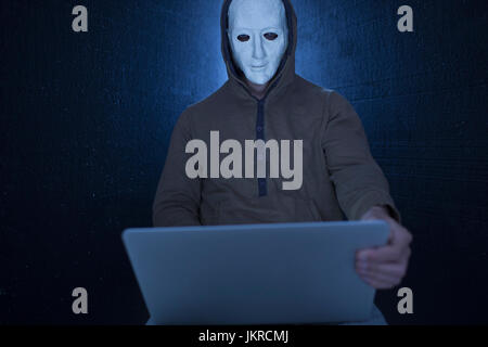 Computer hacker wearing mask using laptop against black background Stock Photo
