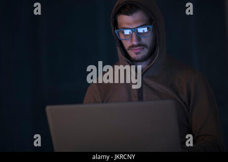 Computer hacker wearing hooded shirt using laptop against wall Stock Photo