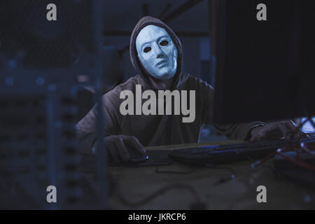 Computer hacker wearing mask and hood using computer while sitting at table Stock Photo