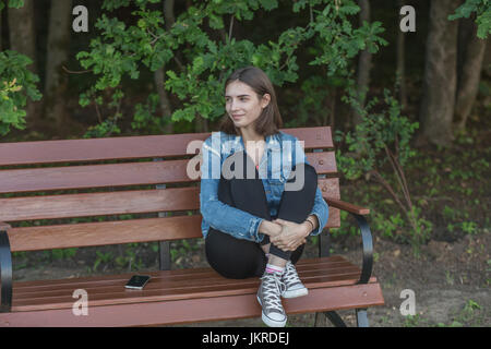 Smiling young woman sitting on wooden bench against trees at park Stock Photo