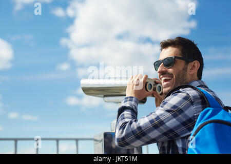 Portrait of young handsome man smiling happily while standing on rooftop against clear blue sky and using coin operated binoculars on viewing platform Stock Photo