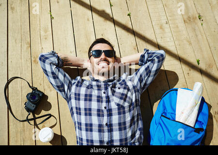 Top view portrait of handsome young man lying on wooden dock planks enjoying sunlight on tourist trip with photo camera and backpack nearby Stock Photo