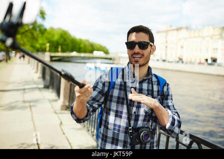 Portrait of handsome young man wearing sunglasses taking selfie photo using smartphone and monopod  on river bank enjoying tourist trip on vacation Stock Photo
