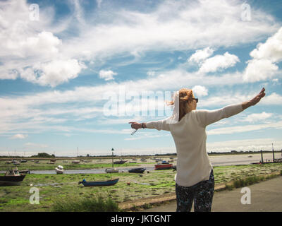 Rear shot of woman in white shirt gesturing with open arms at harbour view of small boats, on a cloudy summer's day in England. Space for text. Stock Photo