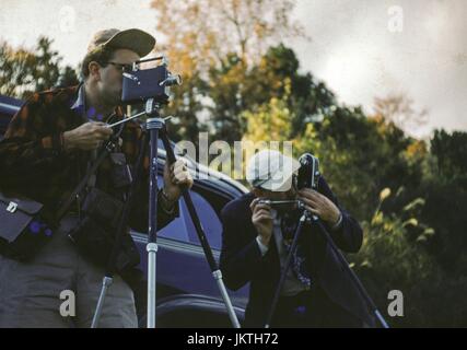 Two men with tripods, both wearing baseball hats, peer through the lenses of 8mm home movie cameras, with a car in the background, Japan, 1952. Stock Photo