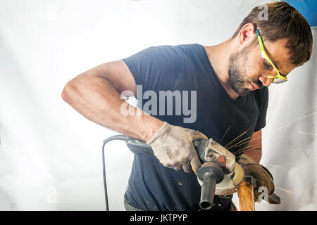 A young man welder in a black T-shirt, goggles and construction gloves processes metal an angle grinder on white isolated backgroun Stock Photo