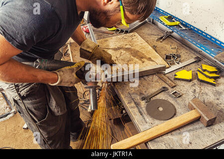 A young man welder in a black T-shirt, glasses and construction gloves  grinder metal an angle grinder   in the workshop, in the background a lot of t Stock Photo