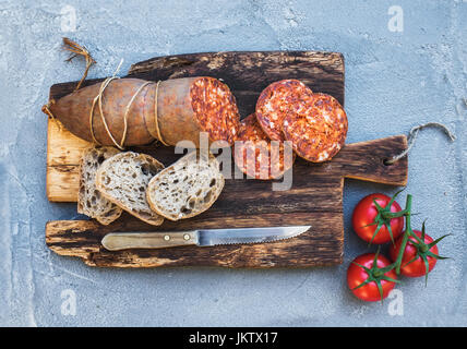 Wine snack set. Hungarian mangalica pork salami sausage, rustic bread and fresh tomatoes on dark wooden board over a rough grey-blue concrete background Stock Photo