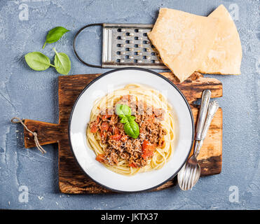 Pasta dinner. Spaghetti Bolognese in metal plate on rustic wooden board with Parmesan cheese, grater and fresh basil over grey concrete background Stock Photo