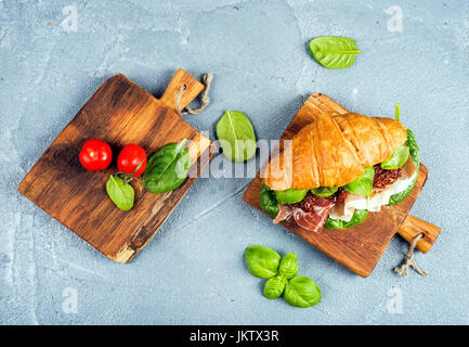 Croissant sandwich with smoked meat Prosciutto di Parma, sun dried tomatoes, fresh spinach and basil on small rustic wooden boards over stone textured grey background Stock Photo