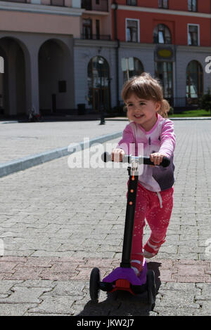 Smiling positive little girl riding on scooter in city Stock Photo