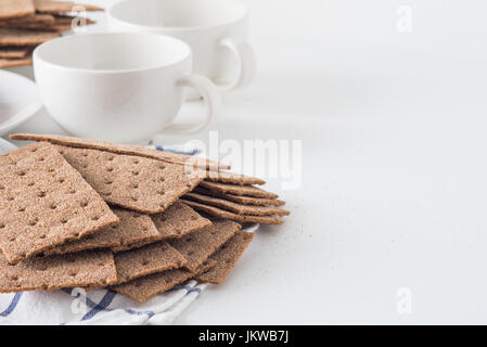 Stack of brown rye crispy bread (Swedish crackers) with two cups and piece of cloth on white background with space for text. Healthy meal concept Stock Photo
