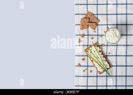 Brown rye crispy bread (Swedish crackers) with spread cottage cheese, decorated with thin green onion, on piece of cloth on light blue background with Stock Photo