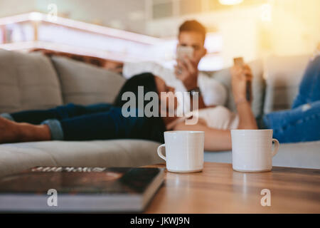 Two coffee cups on table with couple relaxing in background on couch. Cups of coffee in front with man and woman at the back at home. Stock Photo