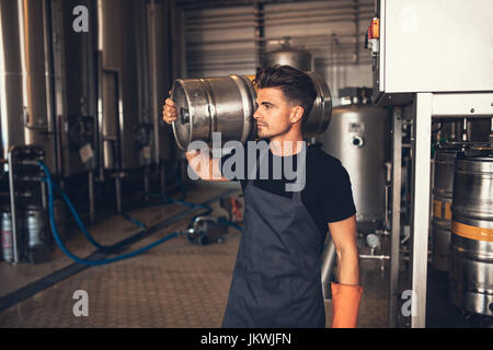 Young male brewer carrying keg at brewery. Manual worker with metal beer barrel craft beer manufacturing plant. Stock Photo