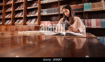 Beautiful young student using tablet pc while writing notes in the library. College student taking notes from reference book and listening music from  Stock Photo