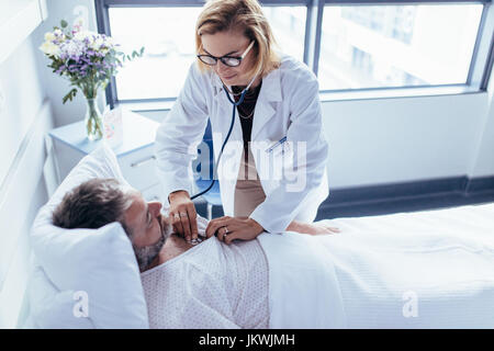 Woman doctor examining heartbeats with stethoscope on a hospitalised man. Physician attending male patient for routine checkup. Stock Photo