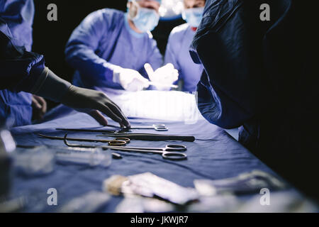 Close-up of nurse taking medical instruments for operation with colleagues doing surgery in background. Medical team performing operation in hospital. Stock Photo