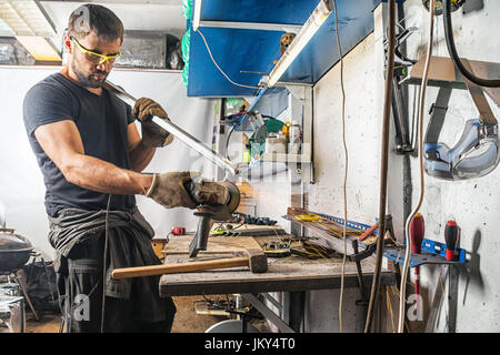 A young man welder in a black T-shirt, goggles and construction gloves grinder metal an angle grinder   in the   workshop,Around a lot of tools for wo Stock Photo