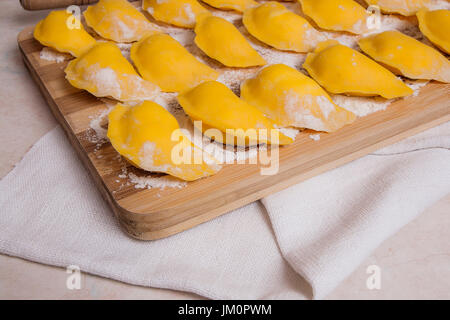 Several ready for boiling vareniki, dumplings, pierogi with cottage cheese or curd on wooden cutting board with flour. Wooden rolling pin, sieve on li Stock Photo