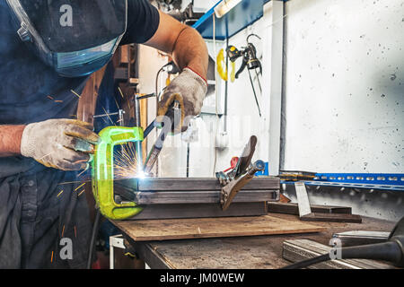 A strong man in a welding mask and working clothes brews a metal angle with a welding machine on a wooden table in the factory, blue sparks fly apart Stock Photo