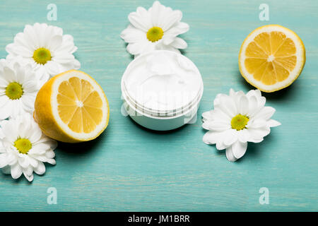 Close-up view of organic cream in container, sliced lemon and chamomiles on blue wooden table Stock Photo