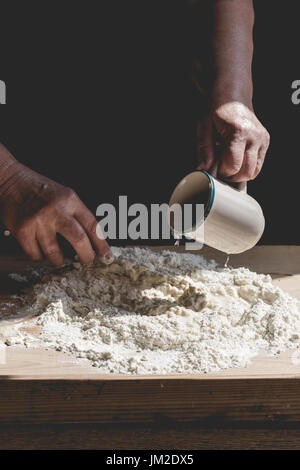 Senior woman kneads pastry, pouring water from mug to flour. Vertical crop, subdued colors, details on old working hands and food ingredients Stock Photo