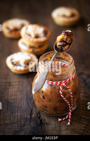 Traditional festive homemade mincemeat in a glass jar and Christmas mince pies at the background on rustic wooden table Stock Photo