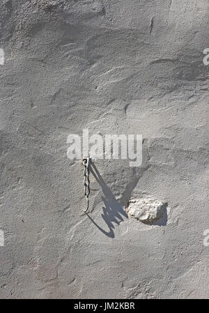 white painted metal chain hanging on textured white rendered wall casting dramatic oblique shadow diagonally on uneven surface in Cumbria, England, UK Stock Photo