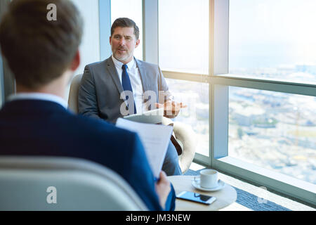 Handome mature corporate manager sitting in a modern office space, holding a digital tablet while interviewing a young business applicant Stock Photo