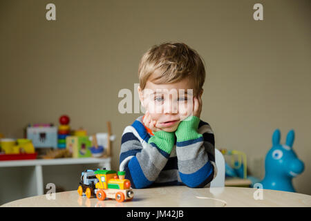A boy toddler sitting in his playroom leans on his elbows on a table and squishes his cheeks with a look of sadness on his face. Stock Photo