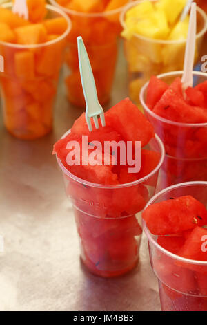 Street food, selection of fruit salads, slices of cut fresh ripe watermelon and melon cubes in plastic cups with forks at retail market stall display, Stock Photo