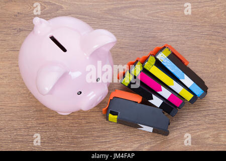 Set of colored and black printer ink cartridges with a pink ceramic piggy bank viewed from above on a wooden desk in a conceptual image Stock Photo