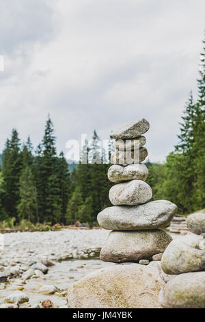 Stones balance, hierarchy stack over cloudy sky in mountains. Inspiring stability concept on rocks, river and forest. Stock Photo