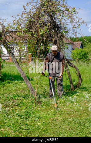local man using a strimmer to cut long grass in the garden of a rural house in a village in zala county hungary