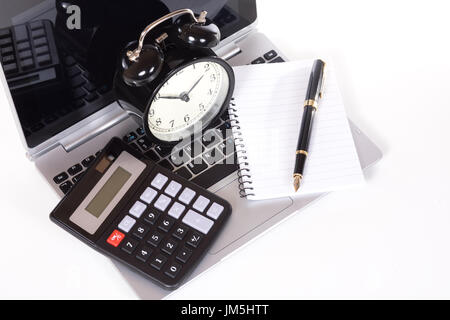 Office objects on laptop keyboard - simple black calculator, blank notebook with fountain pen and classic alarm clock. Viewed from above and isolated  Stock Photo
