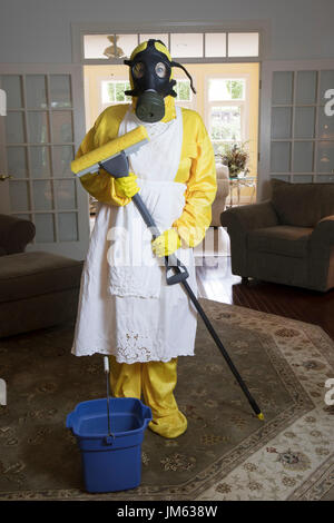 Mature woman in yellow Haz Mat suir and gas mask standing in living room with blue bucket and sponge mop. Stock Photo