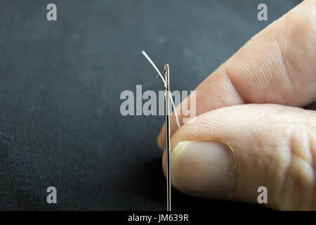 Close up of hand holding white thread in eye of silver needle on black. Stock Photo