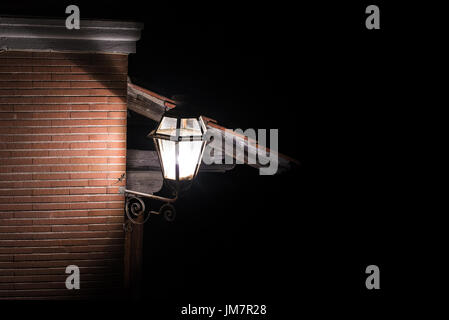 Old fashioned street lamp enlightening brown brick wall and little gecko nocturnal animal on it, dark night background Stock Photo