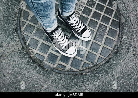 Saint-Petersburg, Russia - May 30, 2017: Teenager feet in a pair of black canvas Chuck Taylor All-Stars casual shoes stand on urban manhole cover