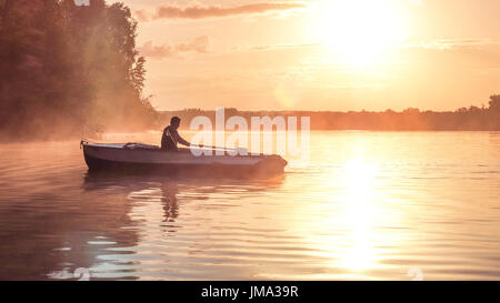 A young guy rides a boat on a lake during a golden sunset. Image of silhouette, sunset. Man rowing a boat in backlight of the sun. Contre-jour, backli Stock Photo