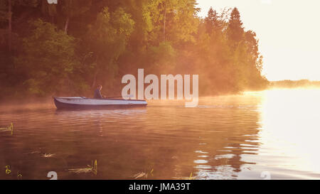 A young guy rides a boat on a lake during a golden sunset. Man rowing a boat in backlight of the sun. Unity with nature concept Stock Photo