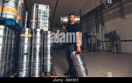 Young male brewer carrying keg at brewery factory. Young man with metal beer barrels at warehouse. Stock Photo