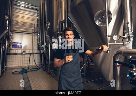 Portrait of male brewer standing by tank in brewery. Man examining the beer in processing section. Stock Photo